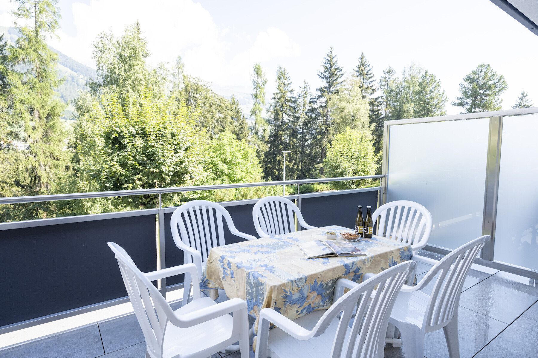 Een balkon met een tafel en stoelen, een metalen hekwerk en uitzicht op bomen en bergen.