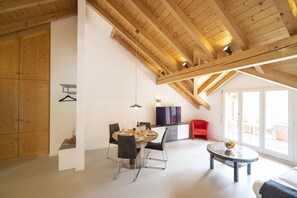 Ein geräumiges Zimmer mit hohen Decken und Holzbalken, einer Essecke mit Tisch und Stühlen, einem roten Sessel, einem kleinen runden Tisch und einem Fernseher auf einem Schrank.