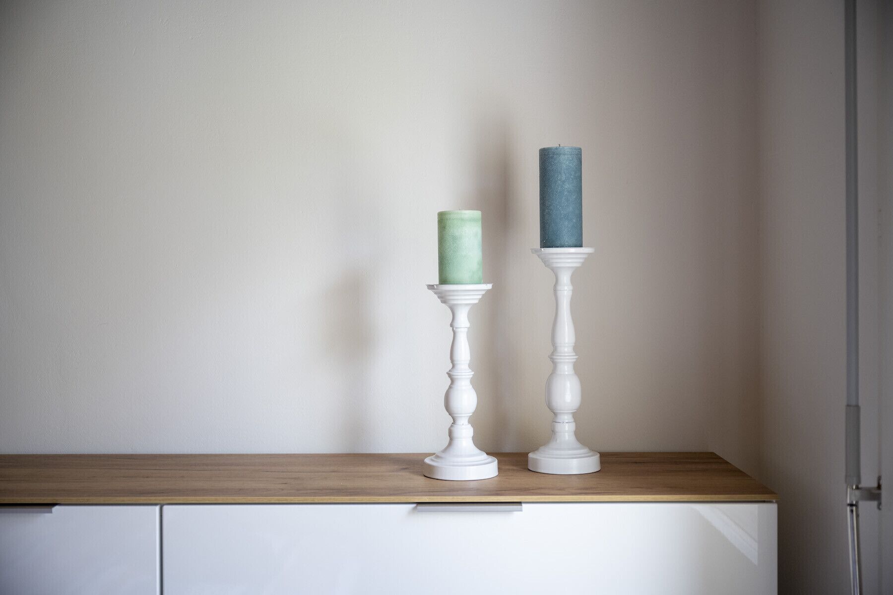 Deux chandeliers blancs, chacun contenant une bougie, sont posés sur une étagère en bois devant un mur uni.