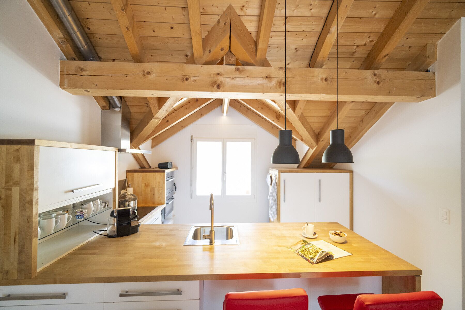 Une cuisine avec des poutres en bois, un îlot central doté d’un évier et deux luminaires suspendus.