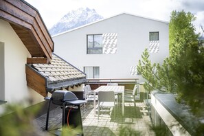 Ein modernes Gebäude mit geneigtem Dach, einem Grillplatz mit Grill, Sitzgelegenheiten im Freien und Blick auf schneebedeckte Berge.