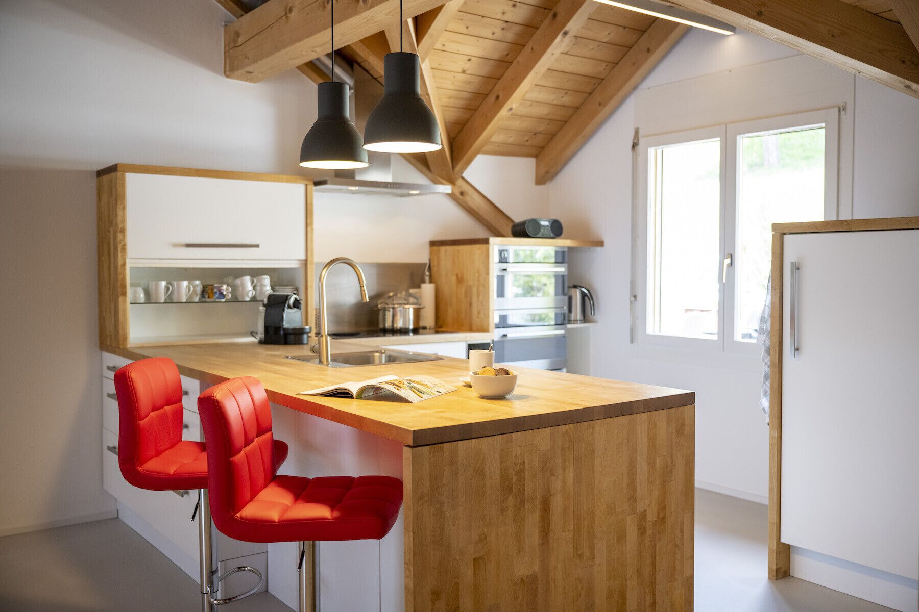Eine moderne Küche mit Holzbalken, einer zentralen Kochinsel mit zwei roten Stühlen und einem großen Fenster für natürliches Licht.