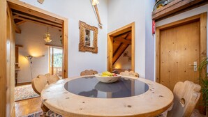 Une table à manger en bois, surmontée d’un plateau en verre, entourée de chaises en bois dotées de coussins à motifs. Un saladier rempli de fruits est posé sur la table.