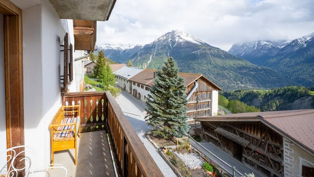 Ein Balkon mit einem Holzstuhl, ein kleiner Garten mit Blumen und Blick auf schneebedeckte Berge.