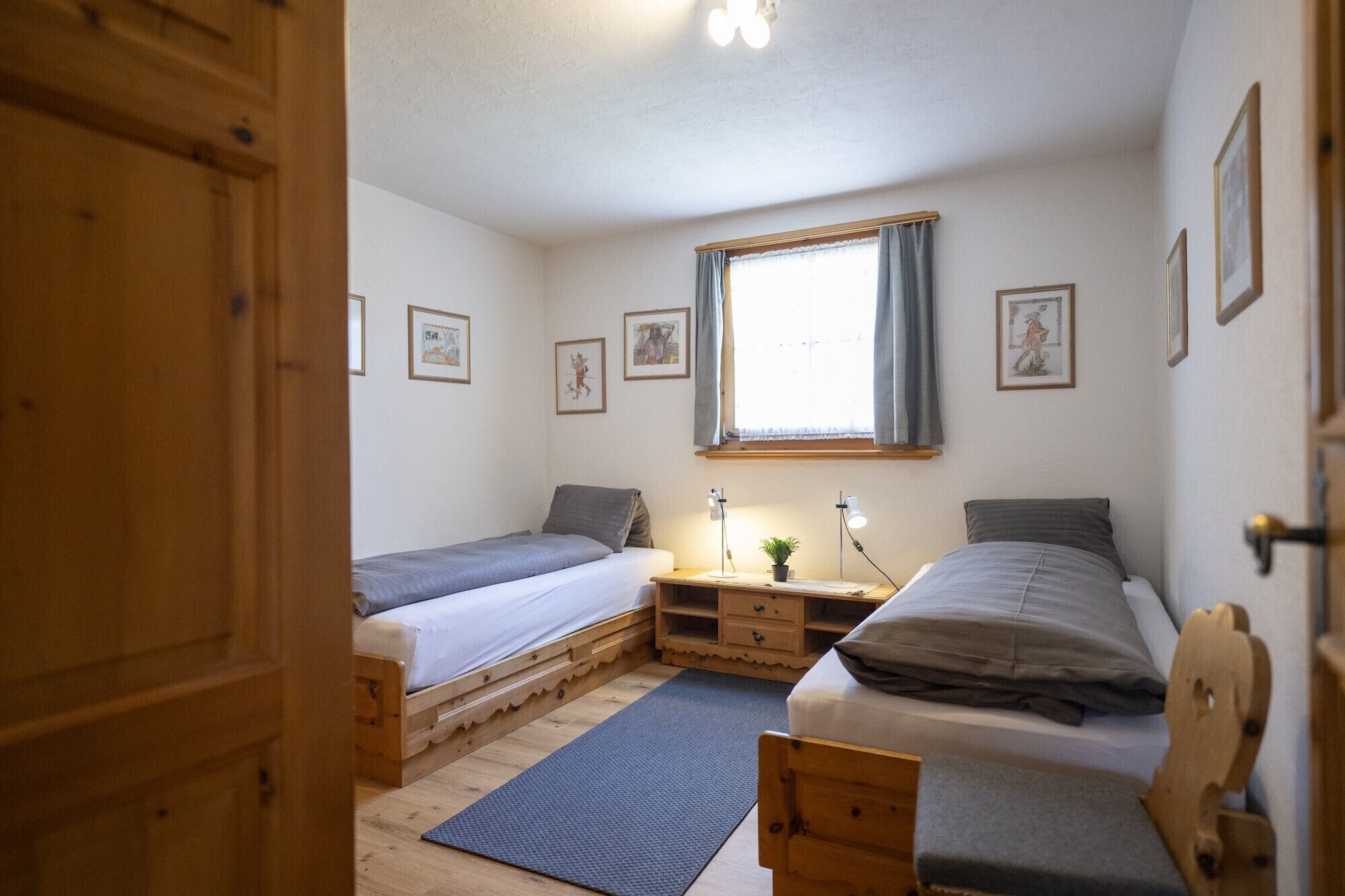 Ein kleines, gemütliches Schlafzimmer mit zwei Einzelbetten, einer Holzkommode, einem Schreibtisch mit Lampe und einem Fenster mit Vorhängen.