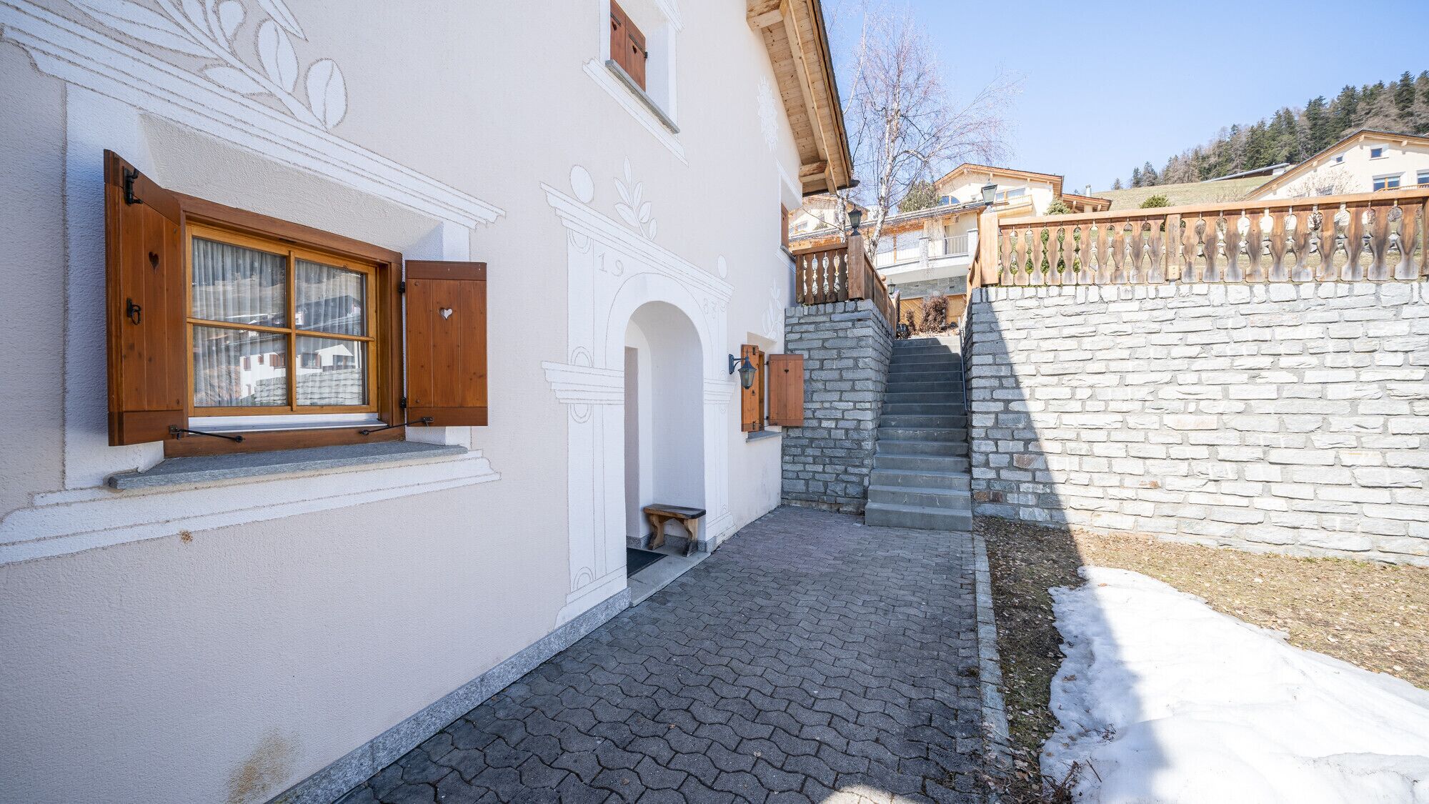 Un bâtiment doté d’un escalier en pierre menant à une terrasse en bois, d’un mur blanc agrémenté d’éléments décoratifs et d’une fenêtre avec des volets en bois.
