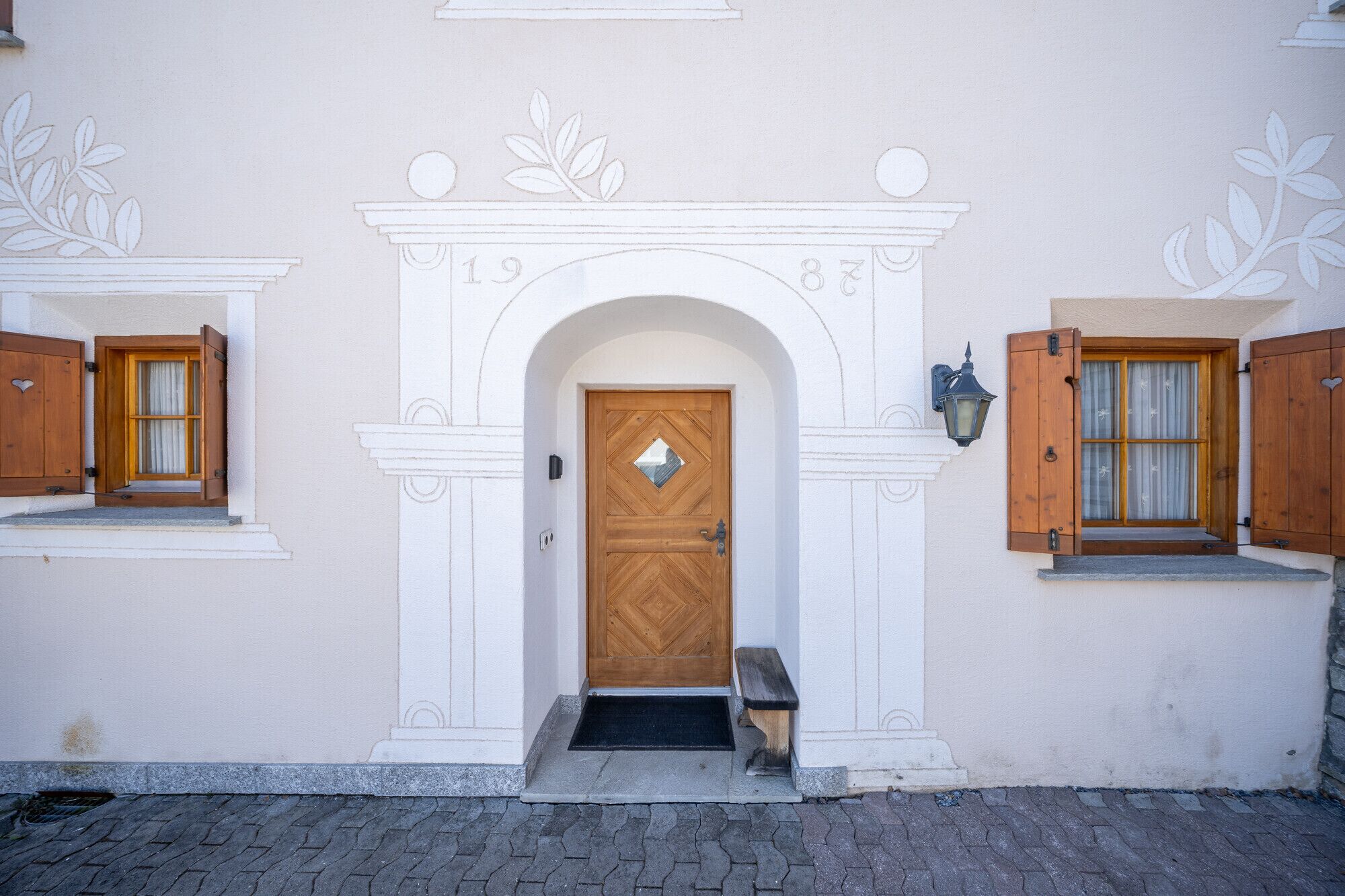 Fachada de edificio con una entrada arqueada central, contraventanas de madera y un pequeño banco en el exterior.