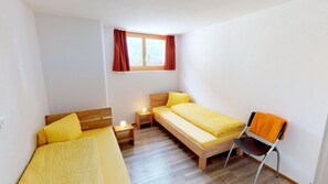 Une petite chambre propre, avec deux lits simples, un plancher en bois, une fenêtre avec des rideaux rouges et une chaise.