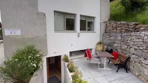 A small, modern building with a stone wall and a patio area. There is a bench and a table with a red cushion. - Chasa Samalgors: Modern and family-friendly 3-room apartment in the idyllic mountain village of Tschlin (Tschlin)