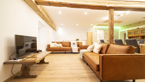 Un salon avec un plafond orné de poutres en bois, un téléviseur posé sur une table en bois, un canapé marron et, au fond, un coin repas.