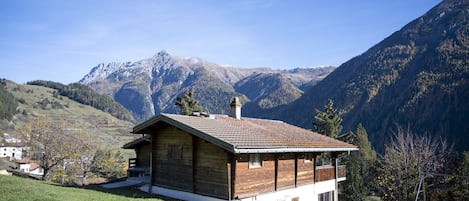 A wooden cabin with a tiled roof, set against a backdrop of mountains and a clear sky.
