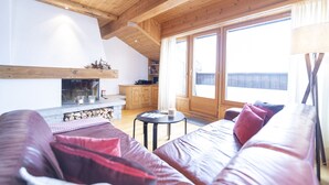 Een gezellige woonkamer met een open haard, houten balken in het plafond, een bank met kussens, een klein rond tafeltje en een schuifdeur naar een balkon.