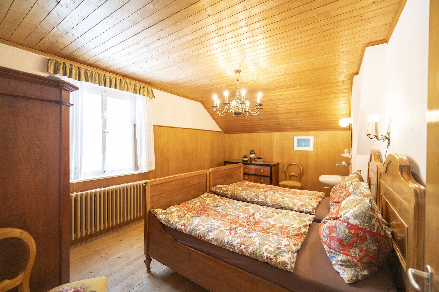 A wooden-paneled room with a bed, a radiator, a window with curtains, a chandelier, and a small table with a vase.