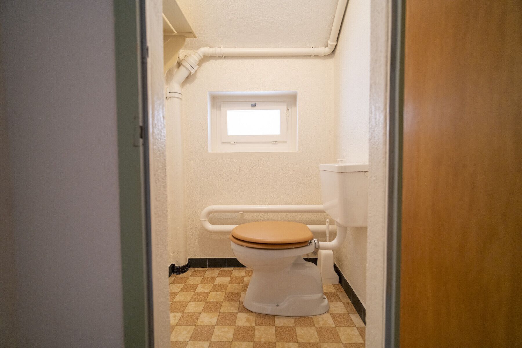 Ein kleines, schmales Badezimmer mit Toilette, Fenster und einem Schachbrettboden.