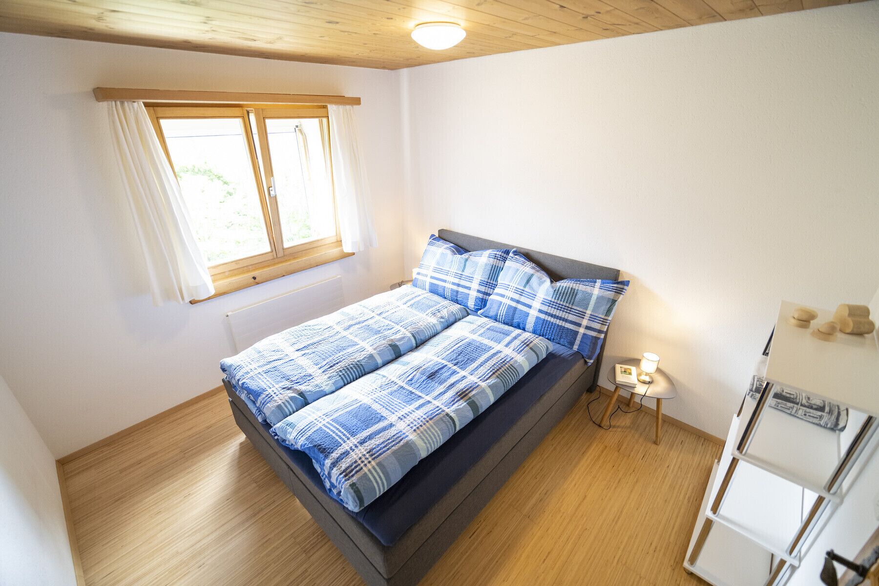 A bedroom with a bed, a window, a bedside table with a lamp, and a white wardrobe.