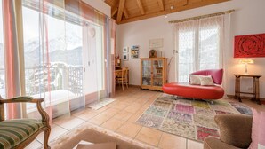 Ein gemütliches Wohnzimmer mit Holzbalkendecke, einem roten Sofa, einer Holzkommode und Blick auf schneebedeckte Berge durch große Fenster.
