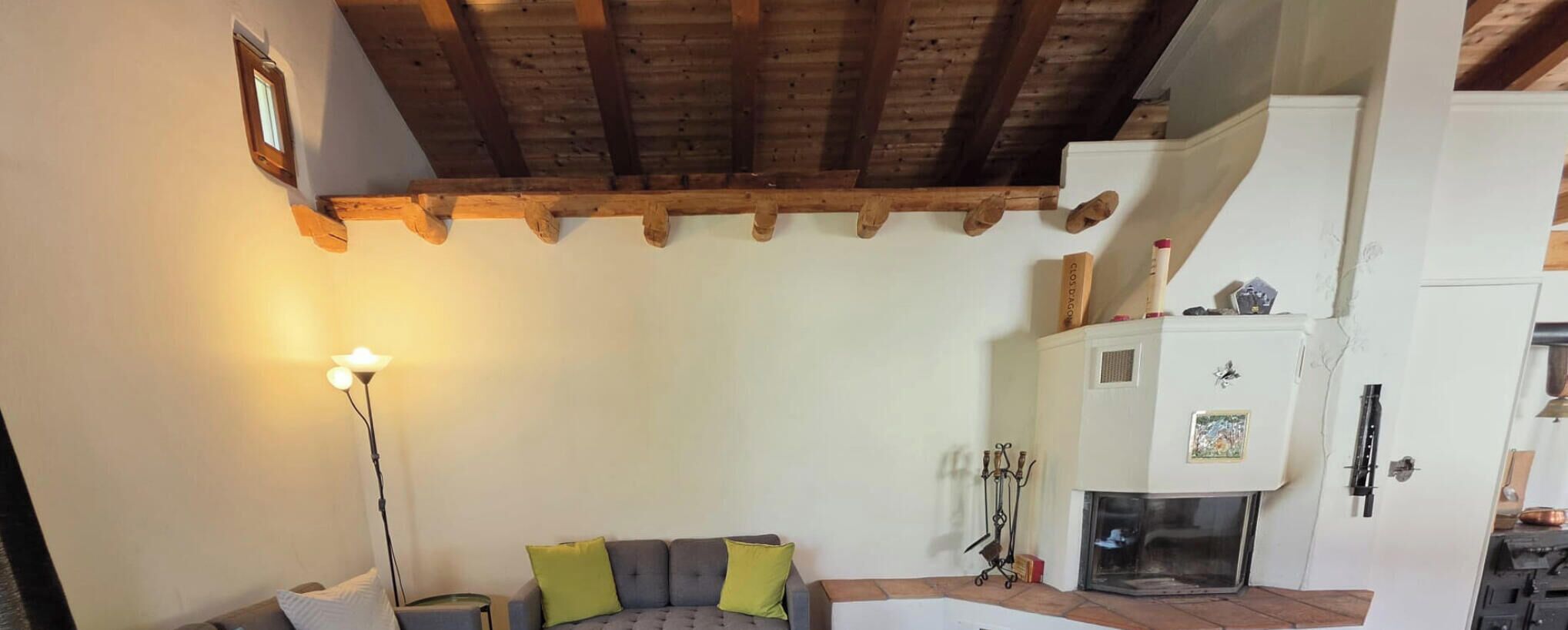 A room with a wooden ceiling, a fireplace, a sofa with cushions, and a floor lamp.