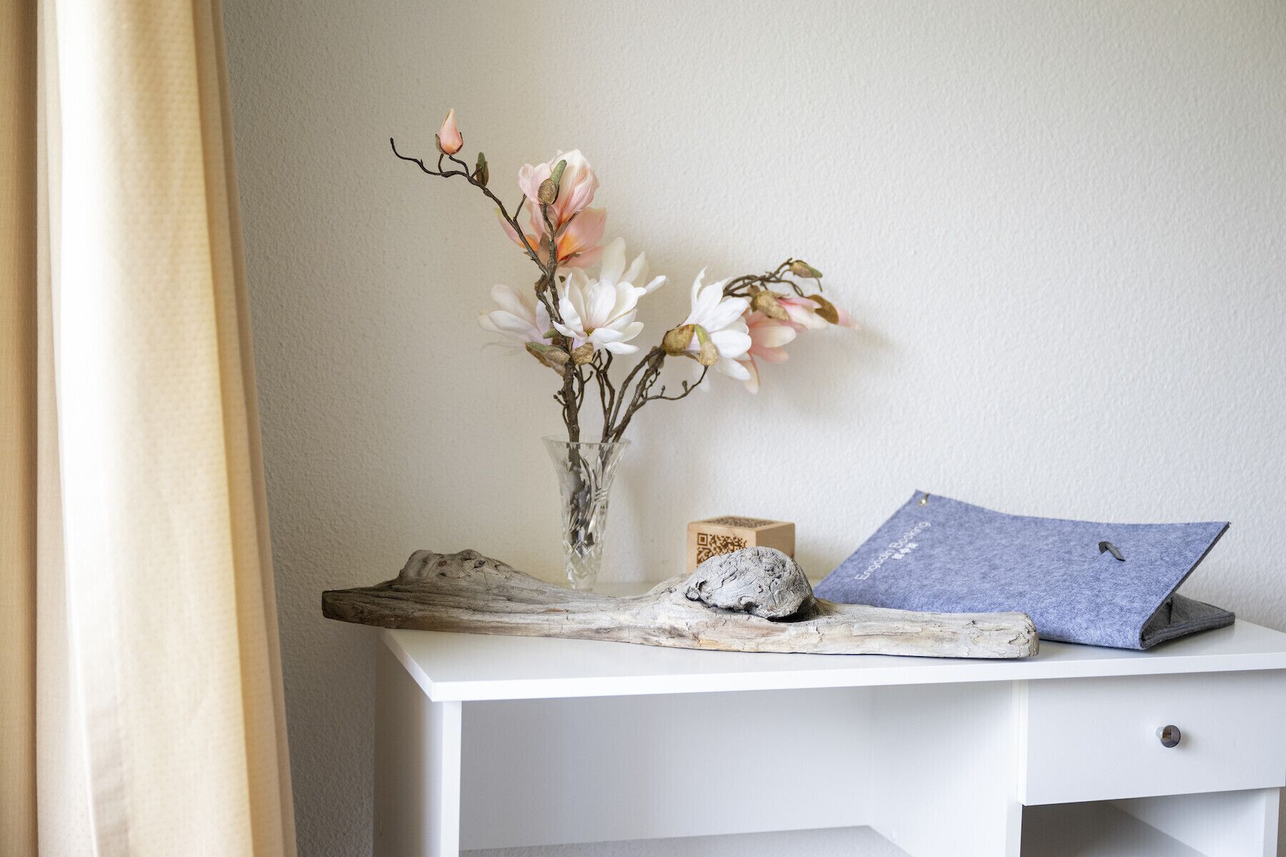 Ein weißer Schreibtisch mit einer Blumenvase, einem Stück Treibholz und einem gefalteten blauen Tuch.