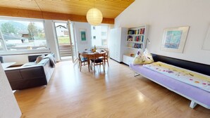 A modern living room with a sofa, a dining table with chairs, a bookshelf, and a large window with a view of the outside. - Chasa Trü No. 7: Trü holiday settlement- Apartment No. 7 (Scuol)