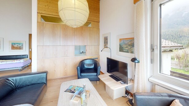 A modern living room with a sofa, a coffee table, a TV, and a view of a forest through a large window. - Chasa Trü No. 7: Trü holiday settlement- Apartment No. 7 (Scuol)