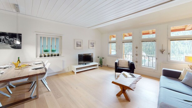 A spacious living room with a wooden dining table, a sofa, and a TV. - Chasa Vista Pisoc: Modern 2 ½-room apartment in the centre with panoramic views (Scuol)
