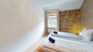 A bedroom with two beds, a stone wall, a window with curtains, and a view of a forest. - Chasa Vista Pisoc: Modern 2 ½-room apartment in the centre with panoramic views (Scuol)