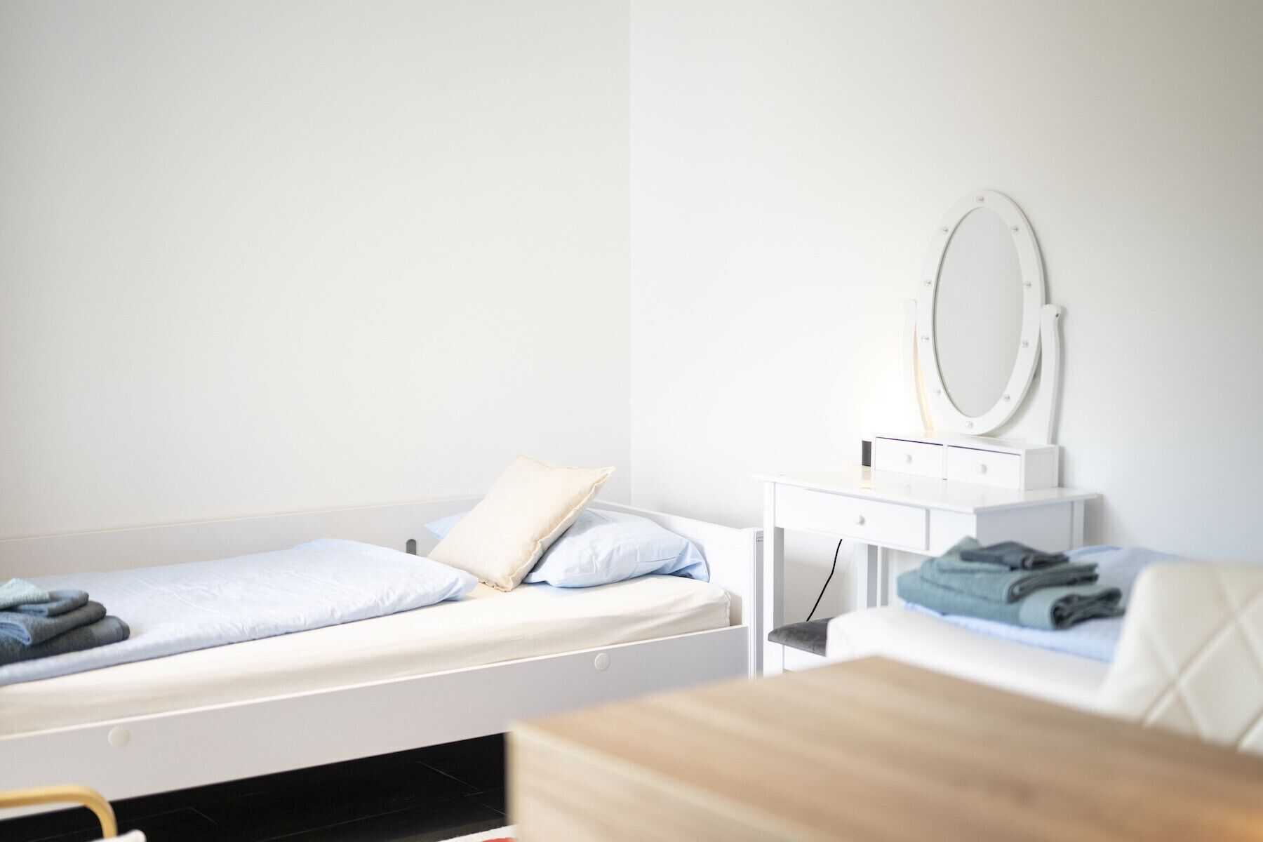 A small, neatly organized bedroom with two beds, a bedside table with a mirror, and a wooden desk.