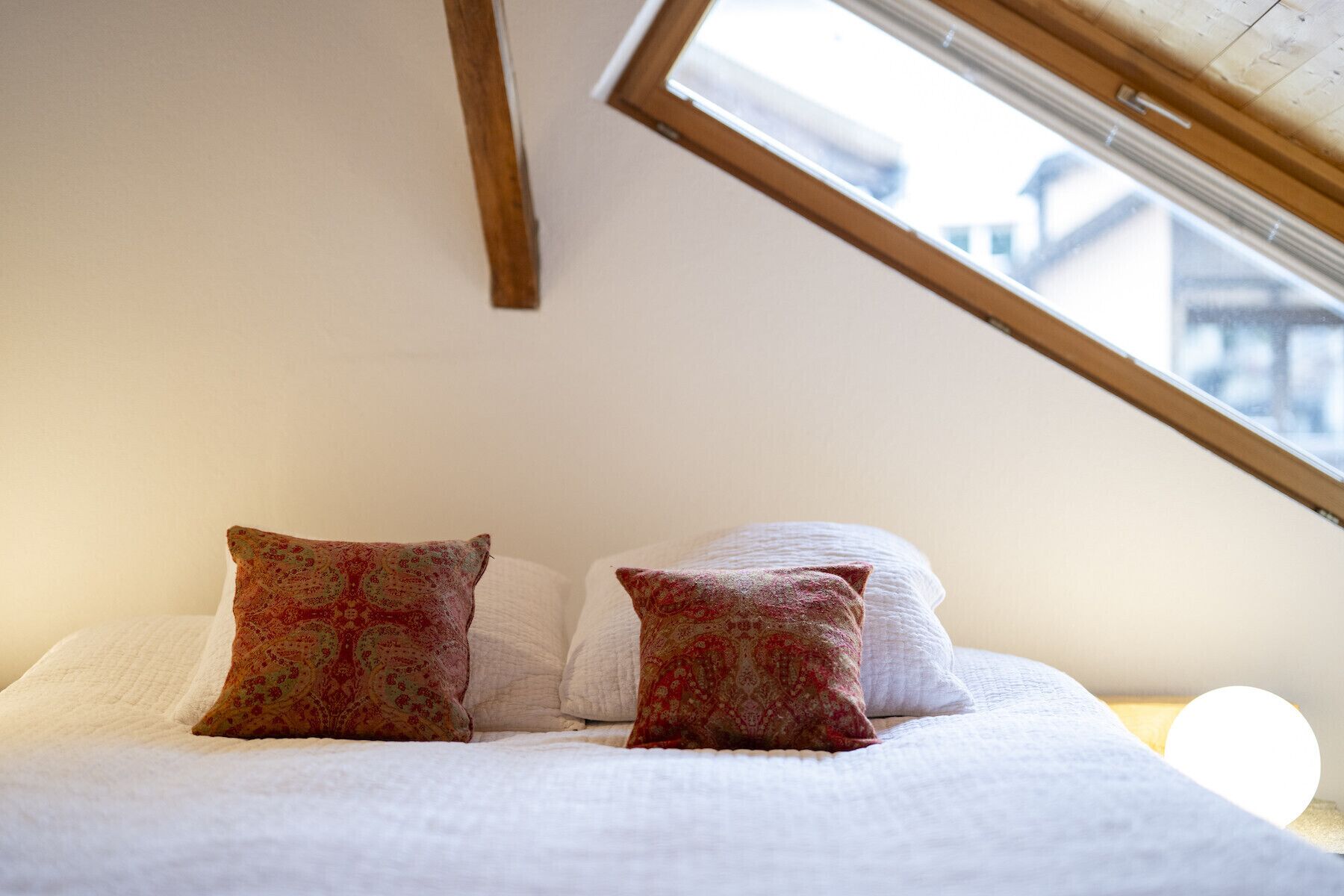 Ein Schlafzimmer mit schräg gedämmter Decke, zwei gemusterten Kissen und Blick durch ein Fenster.