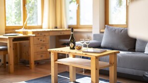 A wooden table with a bottle and glasses, a laptop, and a bowl of snacks in a bright room with large windows. - Chasa Val 735C: Quiet, cozy 3 ½-room apartment with fireplace and panoramic views of the national park (Scuol)