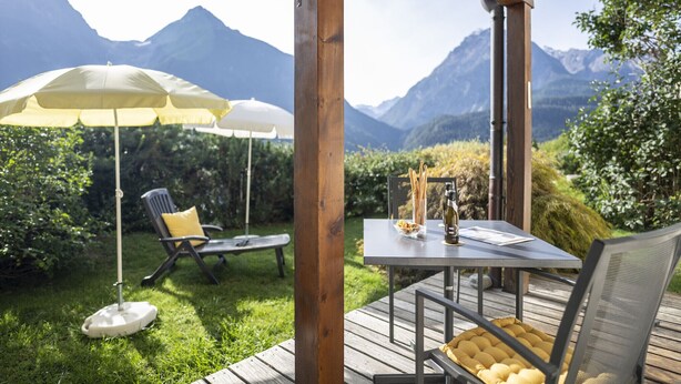 A wooden deck with a table and chairs, surrounded by lush greenery and mountain views. - Chasa Val 735C: Quiet, cozy 3 ½-room apartment with fireplace and panoramic views of the national park (Scuol)