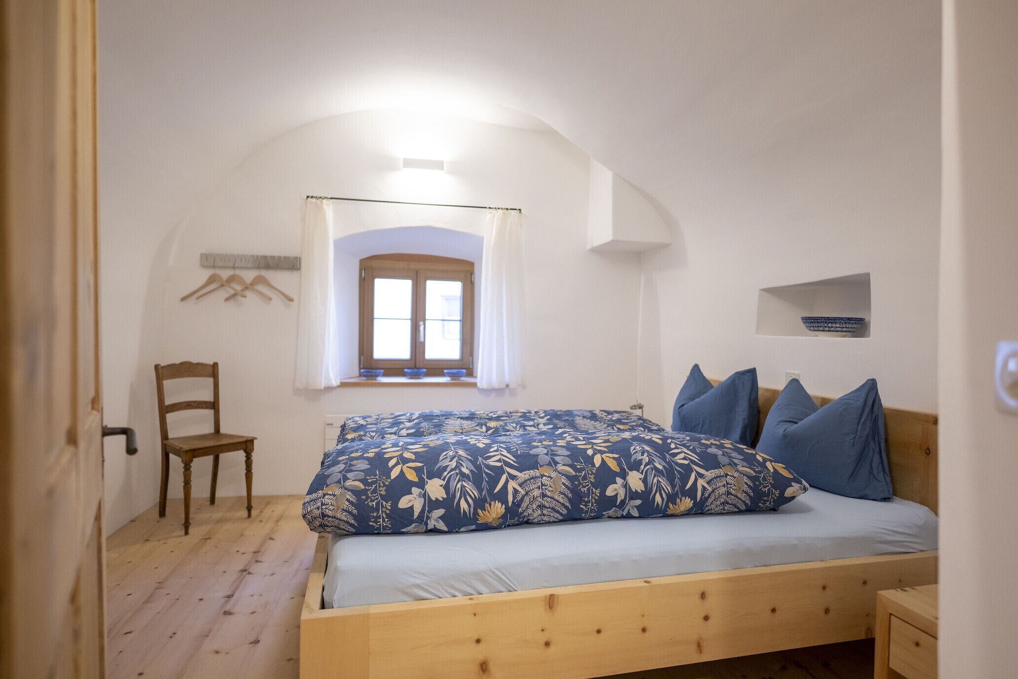 Un dormitorio con una cama de madera, una ventana con cortinas, una silla y un estante pequeño.
