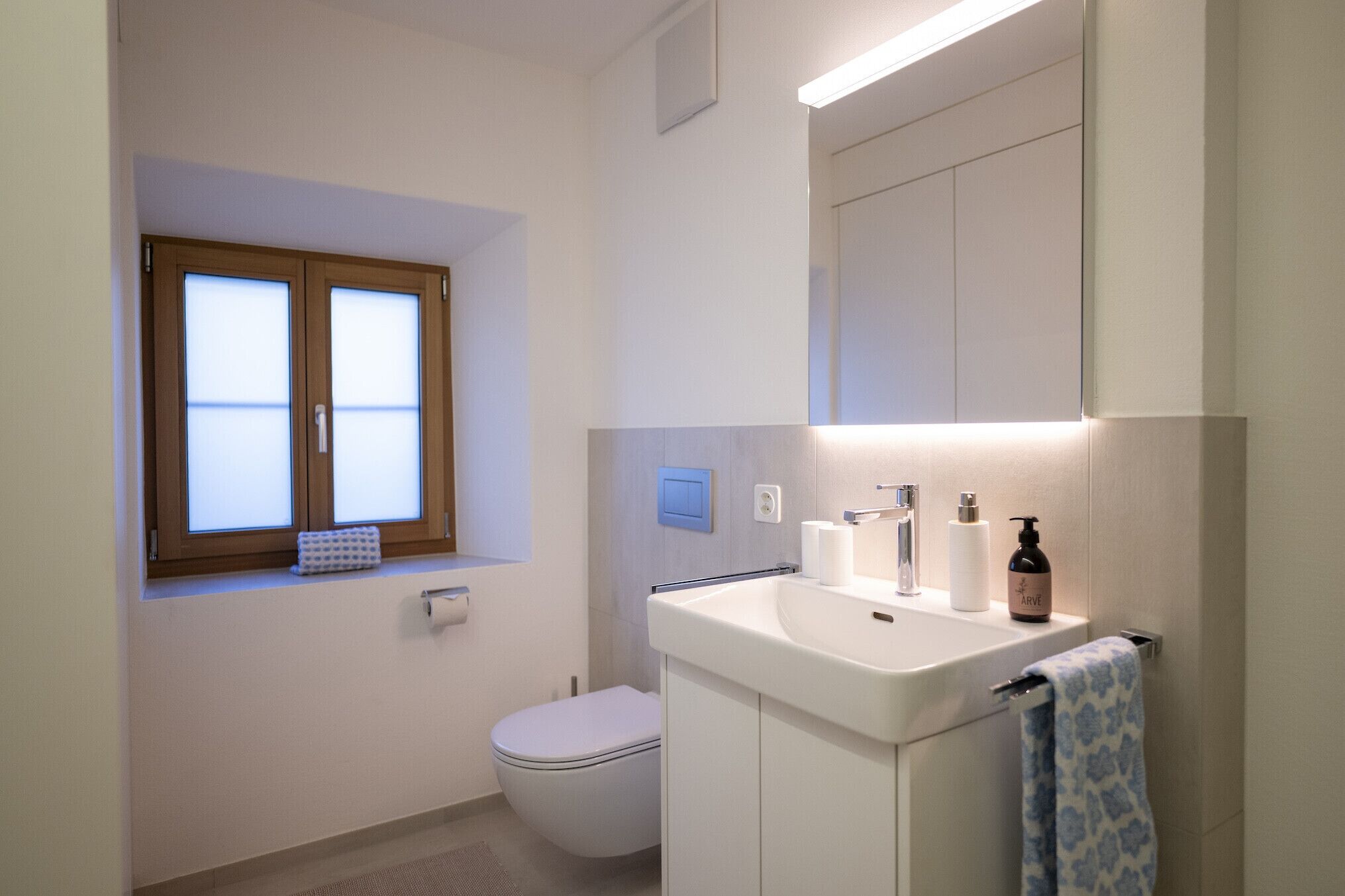 A modern bathroom with a toilet, a sink with a towel, and a window with frosted glass.