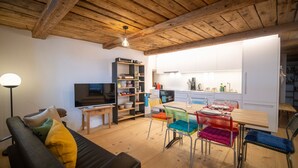 A modern living room with a wooden ceiling, a TV, a bookshelf, and a dining area with colorful chairs. - Chasa Bijou: Idyllic 3-room apartment in the quiet mountain village of Ftan (Ftan)