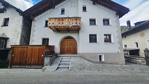 A traditional building with a wooden balcony, a central arched entrance, and a stone staircase leading up to the door. - Chasa Bijou: Idyllic 3-room apartment in the quiet mountain village of Ftan (Ftan)