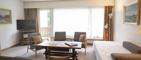 A hotel room with a bed, a sofa, a dining table with chairs, a TV, and a window with curtains.