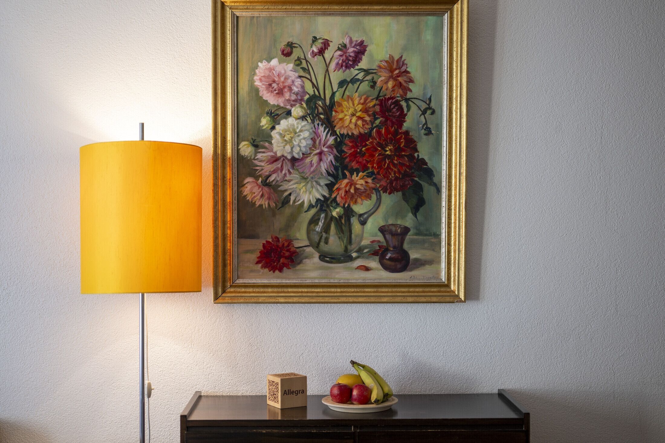 A framed floral painting on a wall, a lamp, a small table with a box labeled "Allegra," and a plate with fruits.
