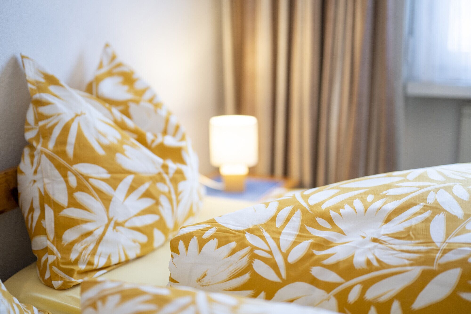 Un llit amb llençols i almoinades de flors grogues i blanques, una làmpada de nit i una finestra amb cortines.
