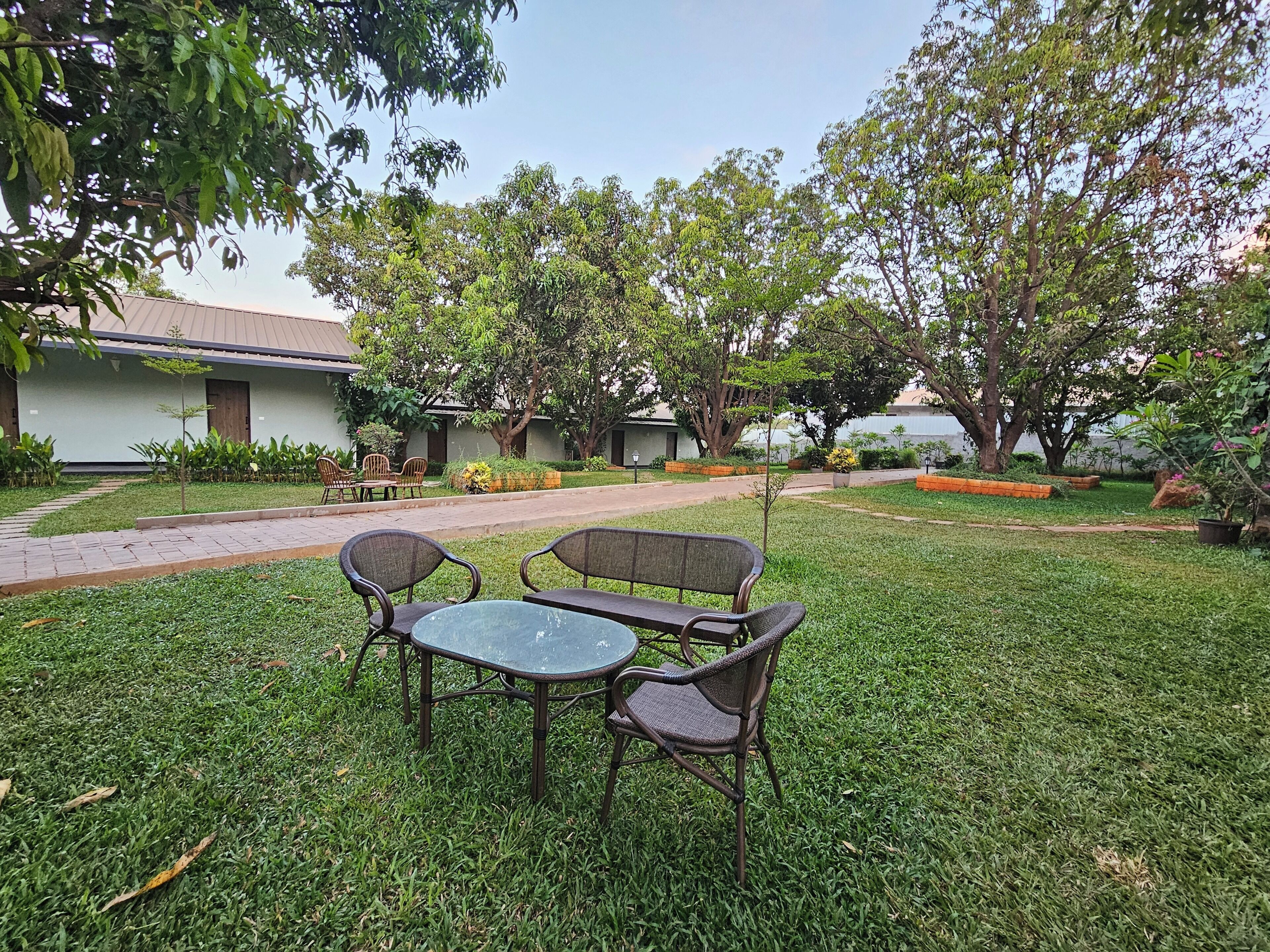 A grassy courtyard with a table and chairs, surrounded by trees and a building with a tiled roof.