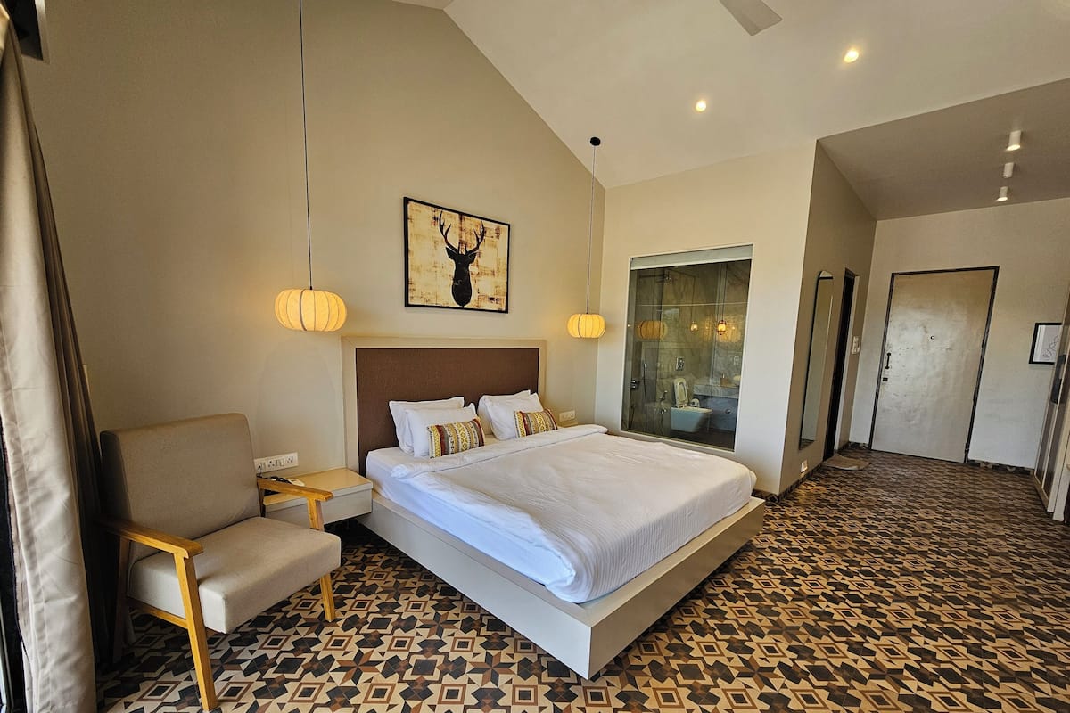 Habitación de hotel con una cama grande, una silla, una mesita de noche, una obra de arte colgada en la pared y una zona de ducha con mampara de vidrio.