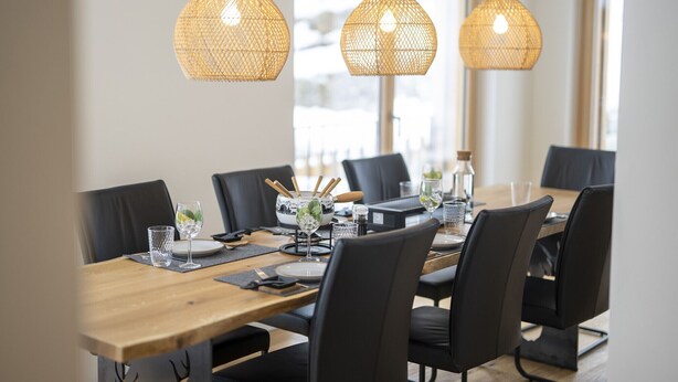 Un comedor con una mesa de madera dispuesta para seis personas, sillas negras y tres lámparas colgantes de mimbre.