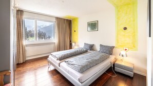 Una camera da letto con una grande finestra, un letto con biancheria grigia, un comodino con una lampada e un quadro appeso al muro.