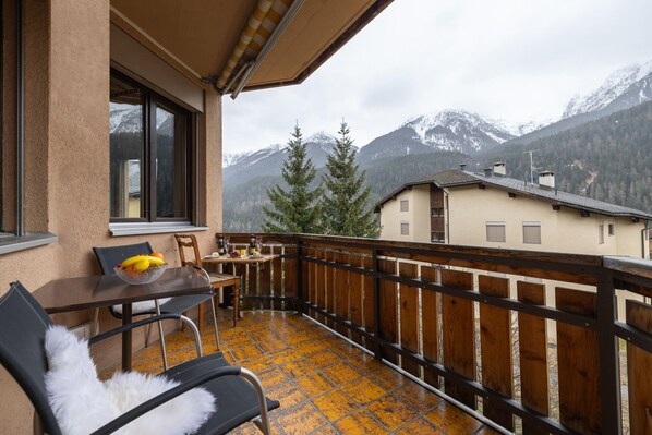 Un balcone con ringhiera in legno, un tavolo con sedie e una finestra con vista sulle montagne innevate.