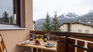 A balcony with a wooden table set for a meal, featuring bread, cheese, and a bottle of wine.
