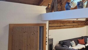 Avokeittiötyylinen huone, jossa on kalteva katto, puupalkit, sohva, sohvapöytä ja pieni keittiöalue, jossa on liesi.