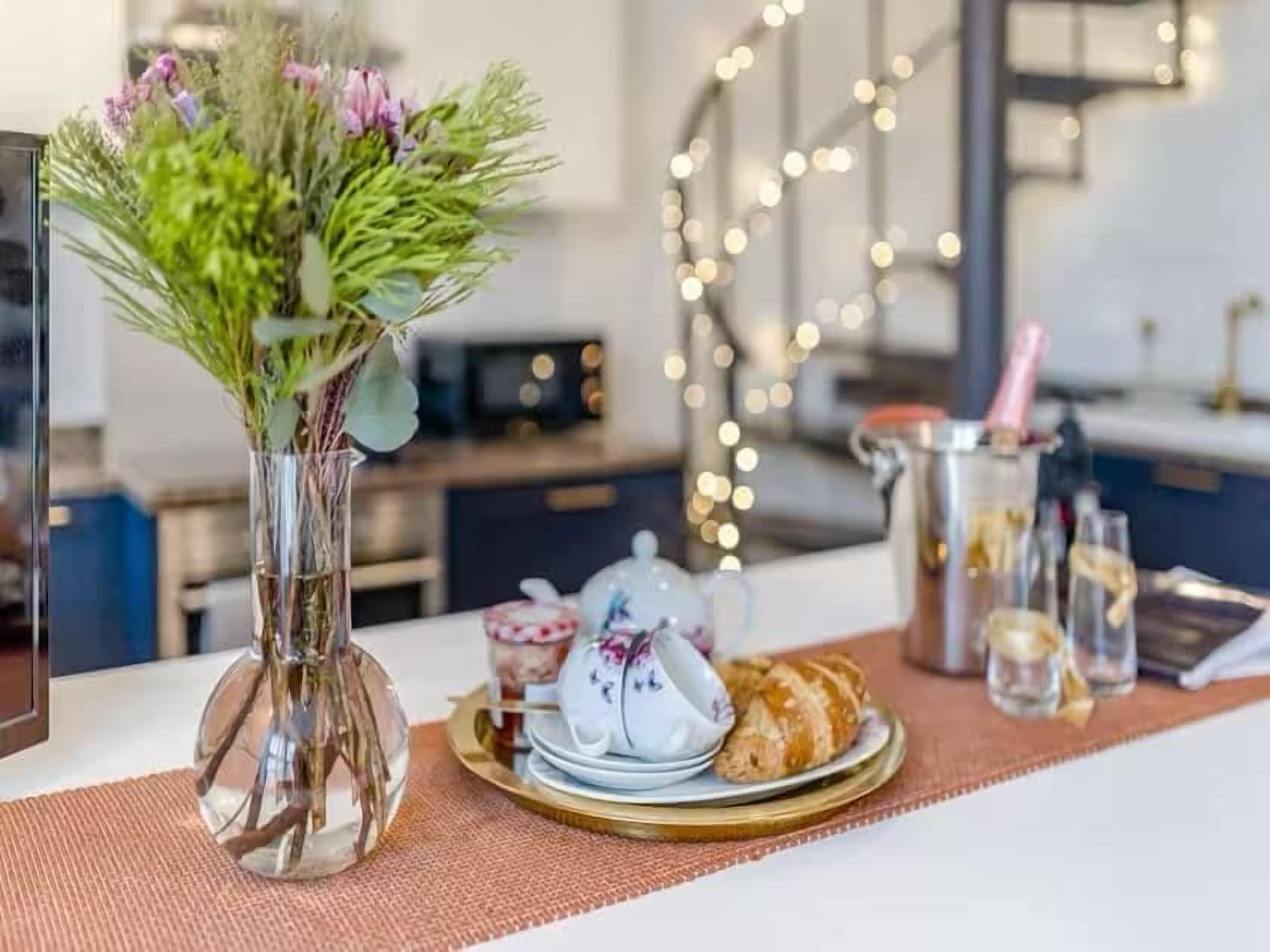 A dining table with a vase of flowers, a teapot, a plate of pastries, and a pitcher of champagne.