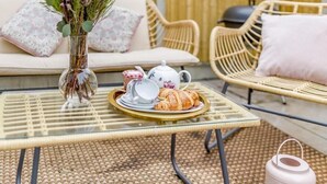 A wicker sofa with cushions, a glass table with a tray of food and tea set, and a vase with flowers. - Railway Cottage - Stylish & Central Oasis in Whitstable (Whitstable)