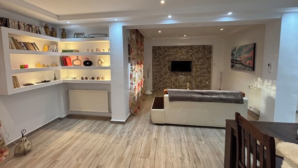 Uma sala de estar com sofá branco, TV fixada em uma parede de pedra e estantes com livros e objetos decorativos.