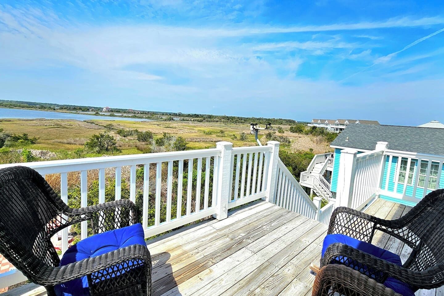 A wooden deck with two wicker chairs, a white railing, and a view of a coastal landscape with buildings in the background.