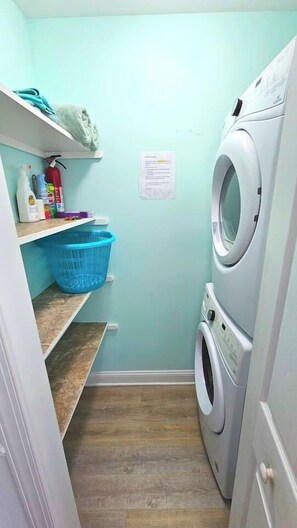 A laundry room with a washing machine, a dryer, a blue laundry basket, shelves with cleaning supplies, and a notice on the wall.