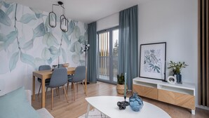 A modern dining area with a wooden table and chairs, a white coffee table, and a wall with a leafy pattern. - RentPlanet - Apartament Prandoty (Kraków)